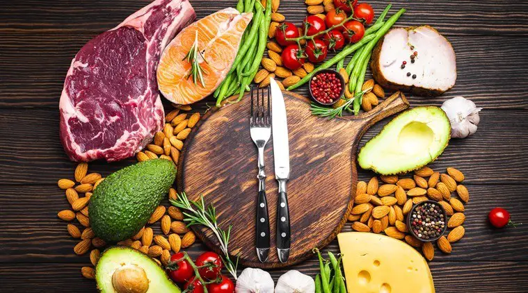 Knife and fork over wooden cutting board and meat, fish, avocado, cheese, vegetables, nuts
